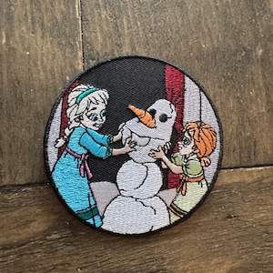 Może przedstawiać: Okrągła naszywka haftowana przedstawiająca dwie dziewczyny budujące bałwana. Dziewczyna po lewej stronie ma na sobie niebieską sukienkę i blond włosy. Dziewczyna po prawej stronie ma na sobie zieloną sukienkę i rude włosy. Bałwan jest biały z marchewkowym nosem i czarnymi oczami.