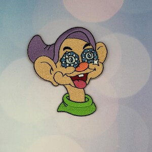 May include: Embroidered patch featuring a cartoon character with a purple hat, large ears, and a green collar. The character has blue and white patterned eyes, an orange nose, and a red tongue sticking out. The patch is likely for clothing or accessories.