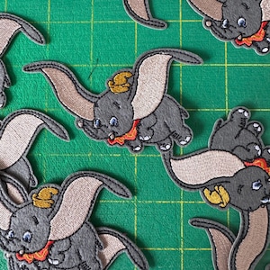 Könnte beinhalten: Gestickte Aufnäher mit der Figur Dumbo, dem Elefanten aus dem Disney-Film. Die Aufnäher sind grau mit beigen Ohren, einem gelben Hut und einem rot-orangen Kragen. Die Aufnäher befinden sich auf einem grünen Hintergrund.