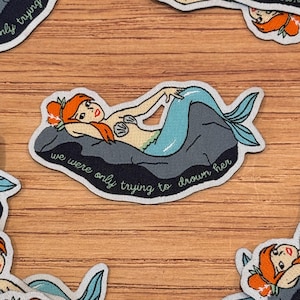 Könnte beinhalten: Ein Stoffaufnäher mit einer Meerjungfrau mit roten Haaren und einem türkisfarbenen Schwanz, die auf einem grauen Felsen liegt. Der Aufnäher hat den Text "we were only trying to drown her" in einer Schreibschrift. Der Aufnäher liegt auf einer Holzoberfläche.
