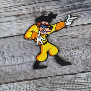 May include: A yellow and black cartoon character wearing sunglasses and a yellow jumpsuit is pointing with one hand. The character is embroidered on a fabric patch.