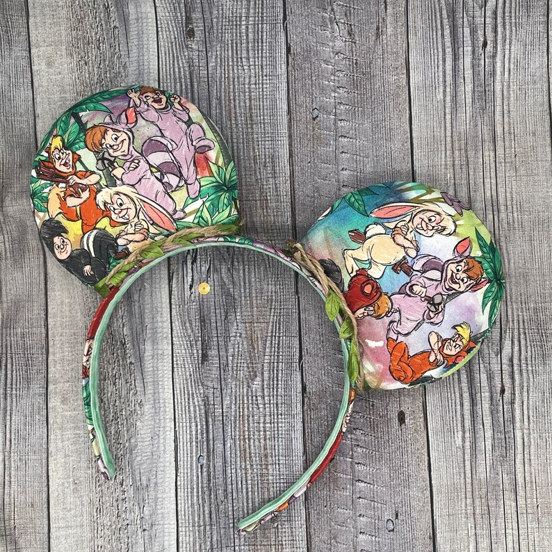 May include: Mouse ears headband featuring a colorful print of characters from a classic animated film. The ears are round and decorated with a jungle-themed fabric. The headband is green and red with a clear plastic center.