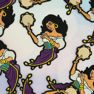 Può includere: Toppa ricamata raffigurante una donna con capelli neri, orecchini a cerchio dorati e un top bianco con spalle scoperte. Indossa una gonna viola con finiture gialle e tiene un tamburello. La toppa ha un design dettagliato a cartoni animati.