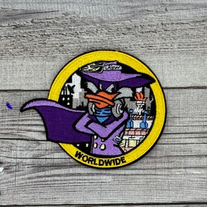 Puede incluir: Un parche bordado con un pato de dibujos animados que lleva un sombrero y una capa morados, con el texto "WORLDWIDE" debajo. El pato sostiene una cámara y tiene un horizonte de ciudad en el fondo.