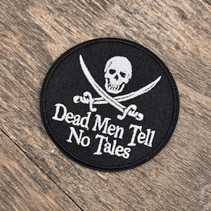 May include: Black circular patch with a white skull and crossed swords design. The text "Dead Men Tell No Tales" is in white, beneath the skull. The patch is on a wooden surface.