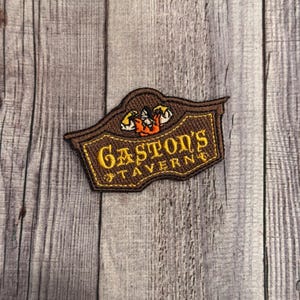 Puede incluir: Parche de fieltro marrón con un letrero bordado en amarillo que dice "Gaston's Tavern". El letrero tiene una imagen de dibujos animados de Gastón de la película de Disney La Bella y la Bestia.