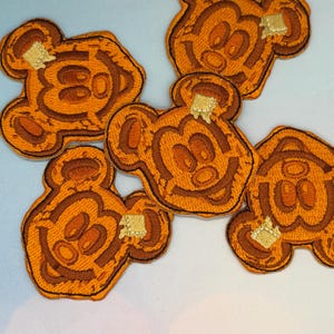 May include: Embroidered patches in the shape of a smiling bear face, resembling a pancake with butter. The patches are primarily orange and brown, with a gold square of butter on each. The patches are likely for clothing or accessories.