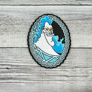 May include: An embroidered iron-on patch featuring Hades from Disney's Hercules. The patch is oval shaped and has a blue and white background with a black and white image of Hades. The patch is surrounded by a black and white decorative border.