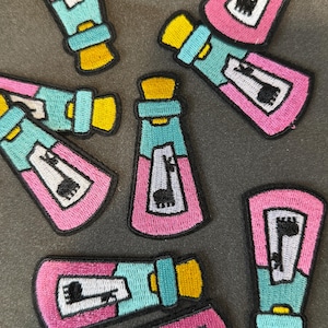 May include: Embroidered patches featuring a potion bottle design. The patches have a pink bottle with a teal top and a yellow cork. The bottle contains a black and white image. The patches are scattered on a gray surface.