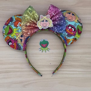 May include: A colorful headband featuring Muppet characters. The headband has large, round ears with a cartoon print of Muppet characters. A rainbow sequined bow is centered on the headband, with a Miss Piggy cutout. A Kermit the Frog sticker is below the bow.