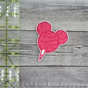 May include: Pink cotton candy shaped like Mickey Mouse ears on a white stick. The cotton candy is embroidered on a white fabric.
