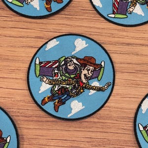 Può includere: Toppa ricamata rotonda raffigurante Woody e Buzz Lightyear che volano in un cielo blu con nuvole bianche. La toppa ha un bordo nero. I personaggi sono in colori vivaci.