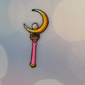 May include: Embroidered patch featuring a pink wand with a gold crescent moon and a white orb at the top. The wand has a detailed design with a star and a bow. The patch is likely for clothing or accessories.