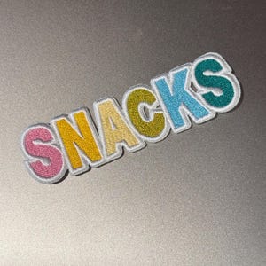 May include: Embroidered patch with the word "SNACKS" in colorful, bubble-style letters. The letters are outlined in white and feature pink, yellow, olive green, and blue hues. The patch is likely for clothing or accessories.