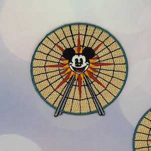 May include: Embroidered patch featuring Mickey Mouse's face at the center of a Ferris wheel design. The patch has a tan background with black lines, red sunburst rays, and a teal border. The Ferris wheel's supports are gray.