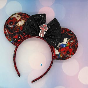 May include: A headband featuring two circular ears covered in a black and red fabric with a Deadpool and unicorn print. A large black sequined bow sits between the ears, adorned with a small, centered Deadpool character charm.