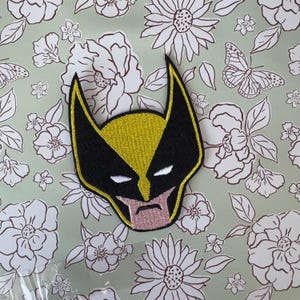 Peut inclure: Écusson brodé représentant le masque d'un super-héros jaune et noir. Le patch a des yeux blancs et une bouche rose. Le fond est vert clair avec un motif floral.
