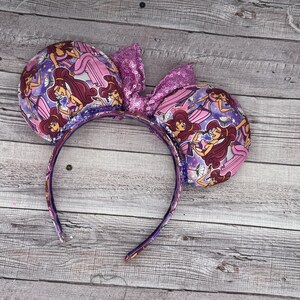 K&ouml;nnte beinhalten: Ein lila Stirnband mit Minnie Mouse Ohren. Die Ohren sind mit einem sich wiederholenden Muster einer Zeichentrickfigur in einem rosa Kleid verziert. Eine paillettenbesetzte Schleife sitzt zwischen den Ohren. Das Stirnband ist lila.
