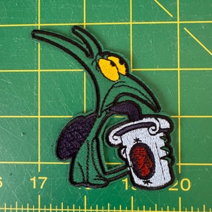 Könnte beinhalten: Grüne Comicfigur mit großen Augen, die eine weiße Tasse mit rotem Logo hält. Die Figur ist auf grünem Stoff gestickt.