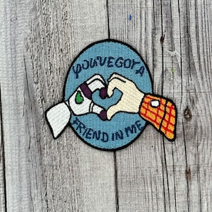 Może przedstawiać: Naszywka haftowana w kolorze niebieskim i białym z tekstem "You've got a friend in me" i dwiema kreskówkowymi dłońmi tworzącymi kształt serca.