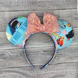 May include: A light blue headband with mouse ears. The ears have a colorful print with text that reads "See Wonder of the World". A large pink sequined bow sits atop the headband. The headband is decorated with pink sequins.