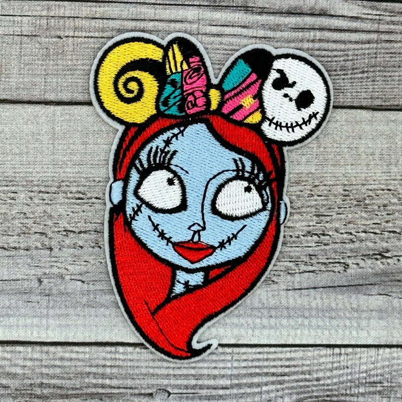 Puede incluir: Parche bordado con Sally de The Nightmare Before Christmas. El parche tiene una cara azul claro, cabello rojo y una apariencia cosida. Una diadema con orejas de Mickey Mouse incluye una cabeza de Jack Skellington y lazos coloridos.