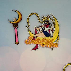 May include: Embroidered patches featuring Sailor Moon and her wand. The Sailor Moon patch shows the character sitting on a crescent moon with the words "Sailor Moon". The wand is pink with gold accents.