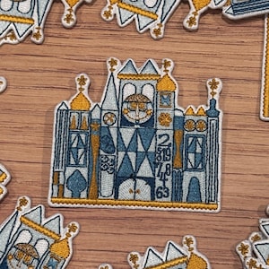 Op de afbeelding: Een geborduurde patch van het iconische kasteel van Assepoester in Walt Disney World. Het kasteel is gedetailleerd met ramen, torens en een klok.