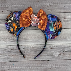 May include: A pair of black headband mouse ears with a sparkly orange bow. The ears are decorated with a space-themed fabric featuring Rocket Raccoon and Groot from Guardians of the Galaxy.