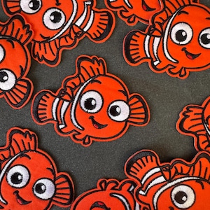 Op de afbeelding: Geborduurde oranje vispatches met witte en zwarte details. De patches hebben een cartoon visontwerp met grote ogen en een lachende mond. De patches zijn gerangschikt op een donkergrijze ondergrond.