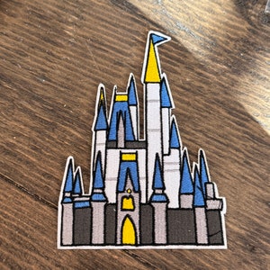 May include: Embroidered patch depicting a stylized castle with blue, white, grey, and yellow details. The castle has multiple turrets and a flag at the top. The patch is outlined in white and is likely for decorative purposes.