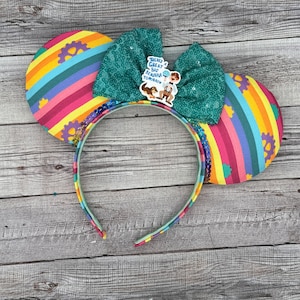 May include: Rainbow striped fabric mouse ears with a green sequin bow and a charm that says "There's a Great Big Beautiful Tomorrow".