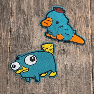 Pode incluir: Dois patches bordados para passar a ferro com personagens de desenhos animados da série de televisão animada Phineas e Ferb. O primeiro patch é de Perry o ornitorrinco, um ornitorrinco azul com um chapéu castanho, um bico laranja e olhos brancos. O segundo patch é de Phineas Flynn, um ornitorrinco azul com um chapéu amarelo, um bico laranja e olhos brancos.