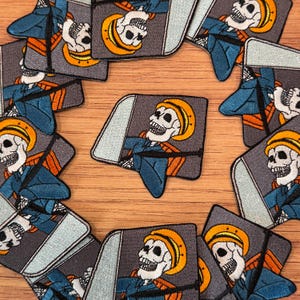 Pode incluir: Uma coleção de patches bordados com um esqueleto usando um chapéu amarelo e uniforme azul, sentado em um veículo. Os patches têm um fundo cinza com uma moldura de janela azul claro e detalhes em laranja. Os patches estão dispostos em um padrão circular.