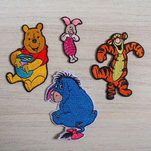 Könnte beinhalten: Ein Set aus vier bestickten Stoffaufnähern mit Winnie Puuh, der einen blauen Honigtopf hält, einem kleinen rosa Ferkel, einem orange-schwarz gestreiften Tigger und einem blauen I-Aah mit einer rosa Schleife am Schwanz, alle mit schwarzen Umrandungen.