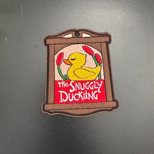 May include: A brown wooden frame with a yellow duckling inside. The frame has the text "The Snuggly Duckling" in red and white lettering.