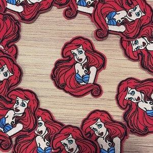 Op de afbeelding: Geborduurde patches met een cartoon zeemeermin met rood haar, blauwe ogen en een blauwe schelp top. De patches zijn rood, wit en blauw en zijn gerangschikt op een lichtbruin houten oppervlak. Voor kleding of accessoires.