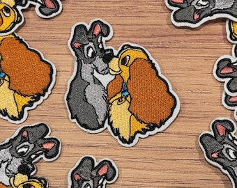 Puede incluir: Parches bordados con los personajes Golfo y Reina de la película animada. Los parches representan a los perros en varias poses, con un perro gris y negro y un perro marrón dorado. Los parches tienen un borde blanco y están sobre una superficie de madera.