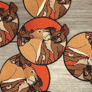 May include: Embroidered patches featuring a cartoon lioness and hyenas. The patches are circular with a red background and black border. The lioness is tan and the hyenas are brown with yellow eyes. The patches are arranged on a light gray surface.