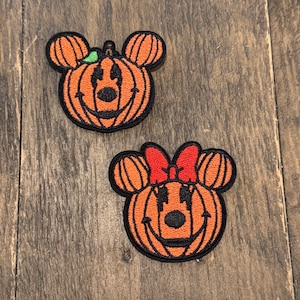 Könnte beinhalten: Zwei Filzpatches von Mickey Mouse und Minnie Mouse, die als Kürbisse verkleidet sind. Die Kürbisse sind orange mit schwarzen Augen und einem schwarzen Lächeln. Mickey Mouse hat einen grünen Stiel auf dem Kopf und Minnie Mouse eine rote Schleife.