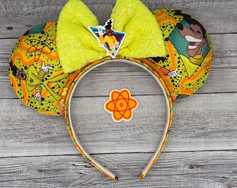 Op de afbeelding: Een hoofdband met een gele paillettenstrik en bijpassende oren. De oren hebben een cartoonontwerp met oranje en gele patronen. Een stripfiguur is te zien op de oren en de strik.
