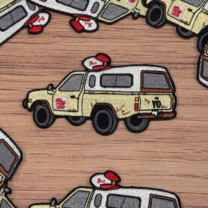 May include: A white and yellow embroidered patch of a pickup truck with a red and white roof light. The truck has a white camper shell and the words "Yo!" on the back. The patch is on a wooden surface.