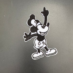 May include: Black and white embroidered patch of Mickey Mouse in a classic pose with his arms raised and legs spread.