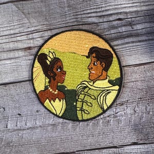 以下が含まれることがあります： 刺繍パッチは、漫画のプリンセスとプリンスの画像を特徴としています。プリンセスは黄色のドレスと王冠を着用しています。プリンスは緑と金の服を着ています。背景は黄色と緑の風景です。