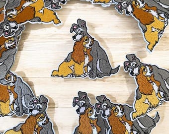 Puede incluir: Parches bordados con los personajes de La Dama y el Vagabundo de la película de Disney. Los parches muestran a los dos perros abrazándose, con La Dama en amarillo y marrón, y el Vagabundo en gris. Los parches están dispuestos sobre una superficie de madera clara.