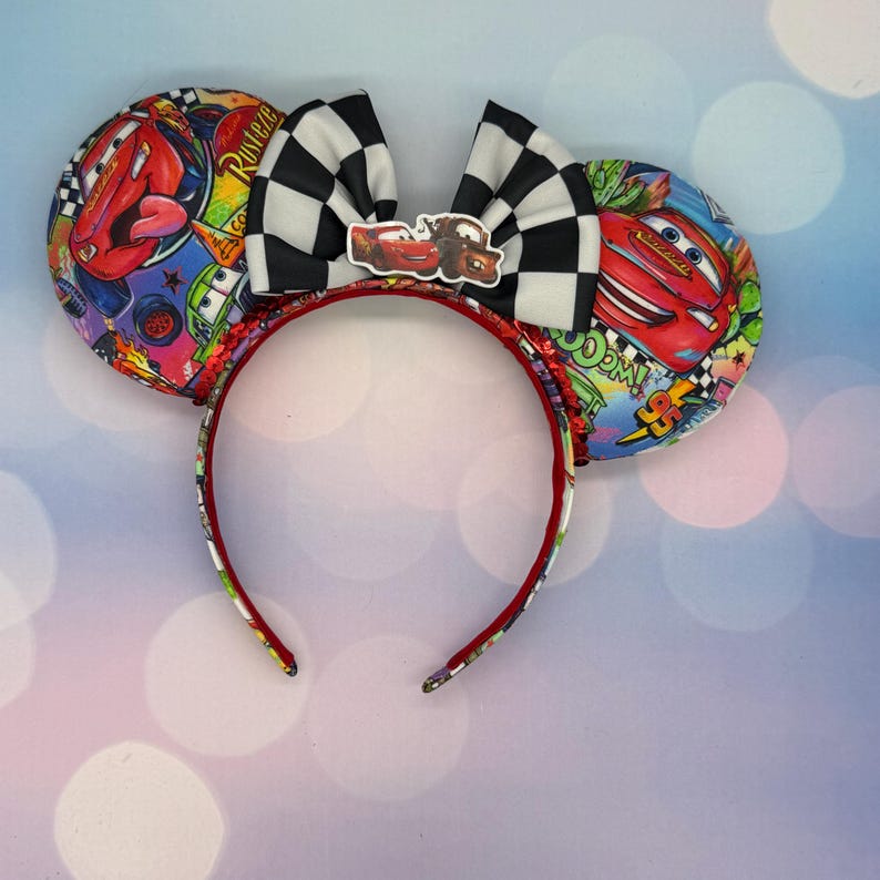 May include: A pair of mouse ears featuring a red headband with Cars-themed ear covers. The ears have a black and white checkered bow with a Cars character emblem in the center. The ear covers display Lightning McQueen and other characters.