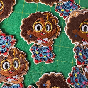 Puede incluir: Parches bordados con un personaje de dibujos animados con piel morena, cabello castaño rizado y gafas. El personaje viste un vestido azul con detalles florales y un lazo rojo. Los parches tienen un borde dorado.