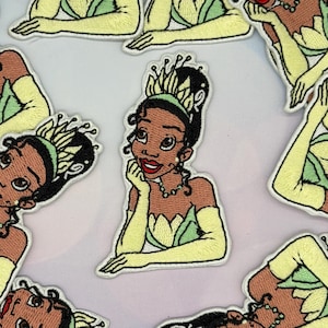 May include: Embroidered patch featuring a cartoon princess with brown skin, wearing a green and yellow dress, gloves, and a crown. The princess is smiling and resting her chin on her hand. The patch has a white border.