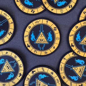 May include: Circular black and yellow embroidered patches with a central triangle eye design, surrounded by symbols and blue flames. The patches are arranged on a dark gray surface.