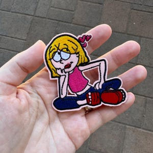 Puede incluir: Parche bordado con una chica de dibujos animados con pelo rubio, que lleva una camiseta rosa, vaqueros azules y zapatos rojos. El parche tiene un borde rosa e incluye pequeños corazones rosas. La chica está en una pose relajada.